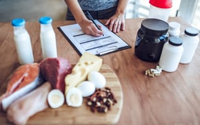A person writes a nutrition plan beside salmon, chicken, eggs, cheese, nuts, protein powder, and supplement bottles