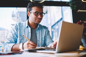 male student studying on laptop, making notes in notepad