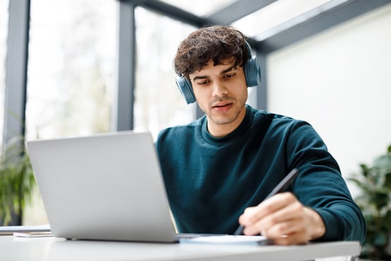 college student in a sweater and headphones, taking notes while studying on his laptop