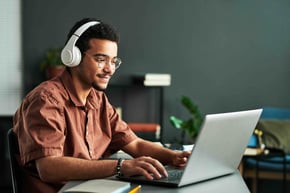 A person wearing headphones and glasses sits at a desk using a laptop, with books and plants in the background.