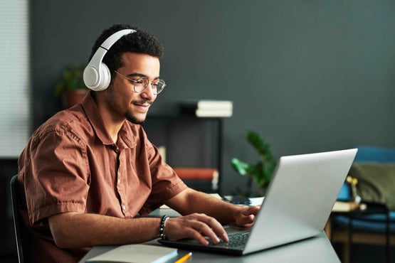 A person wearing headphones and glasses sits at a desk using a laptop, with books and plants in the background.