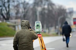 Construction surveyor using total station on tripod to collect geospatial data in city. Land surveyor wearing protective clothing operating geodetic equipment for urban development planning