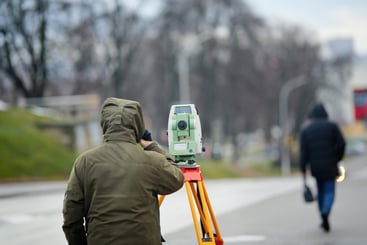 Construction surveyor using total station on tripod to collect geospatial data in city. Land surveyor wearing protective clothing operating geodetic equipment for urban development planning
